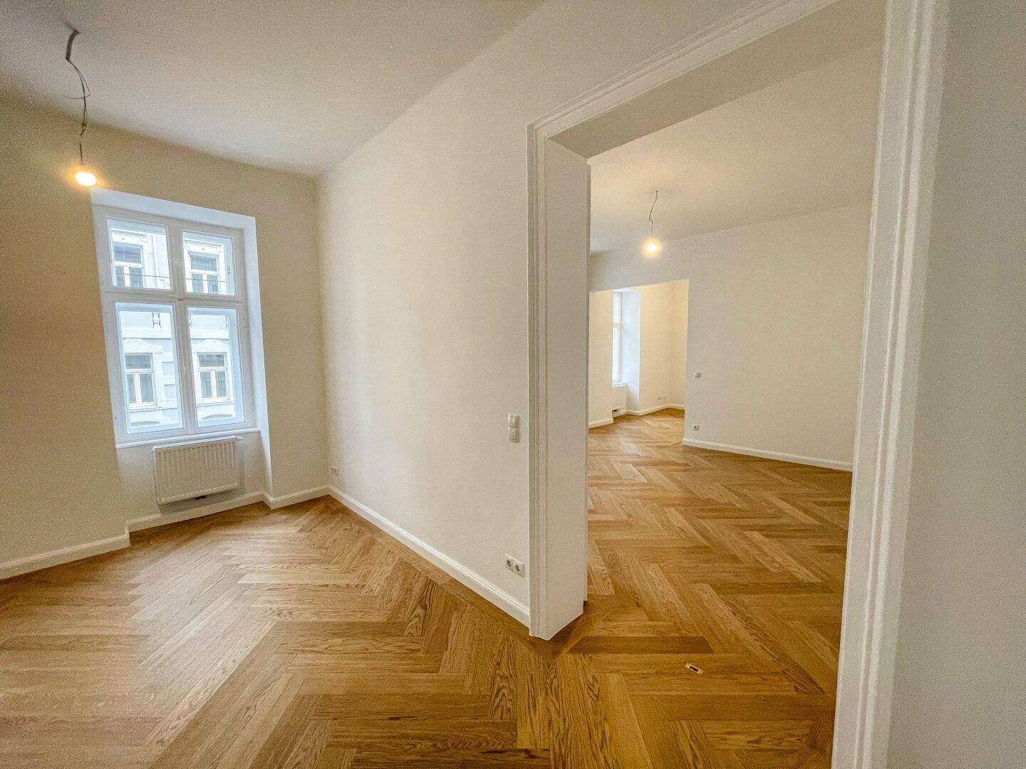 Geräumiges Zimmer mit Fischgrätparkett und offenem Durchgang zu einem weiteren hellen Raum.