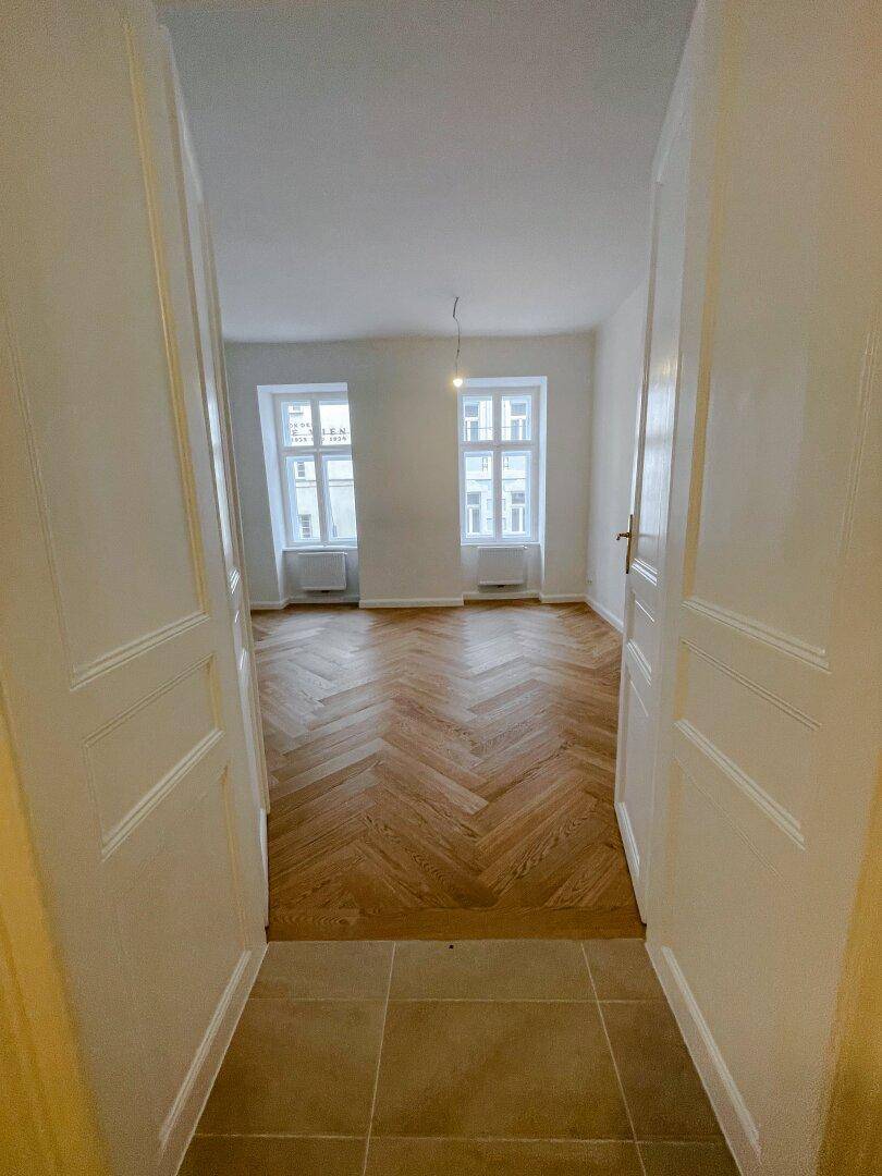 Blick vom gefliesten Flur in ein helles Zimmer mit klassischem Fischgrätparkett und zwei Fenstern.