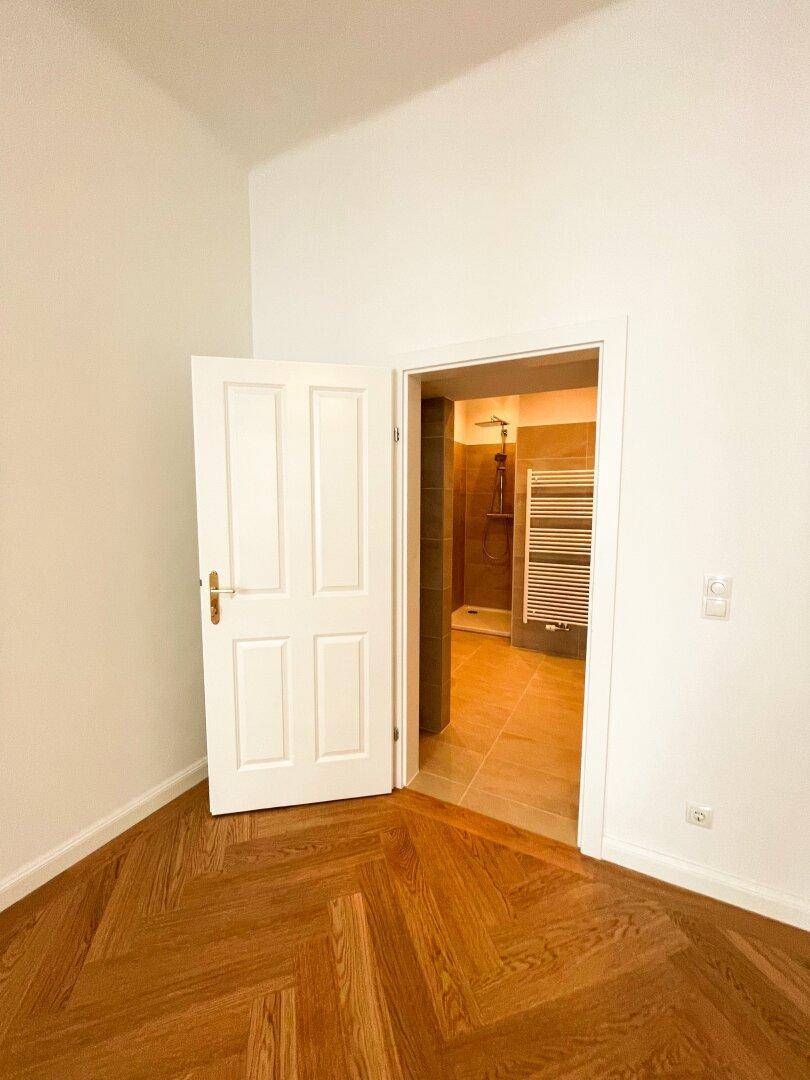 Blick aus einem Zimmer mit Fischgrätparkett in ein modernes Badezimmer mit Dusche und Handtuchheizkörper.