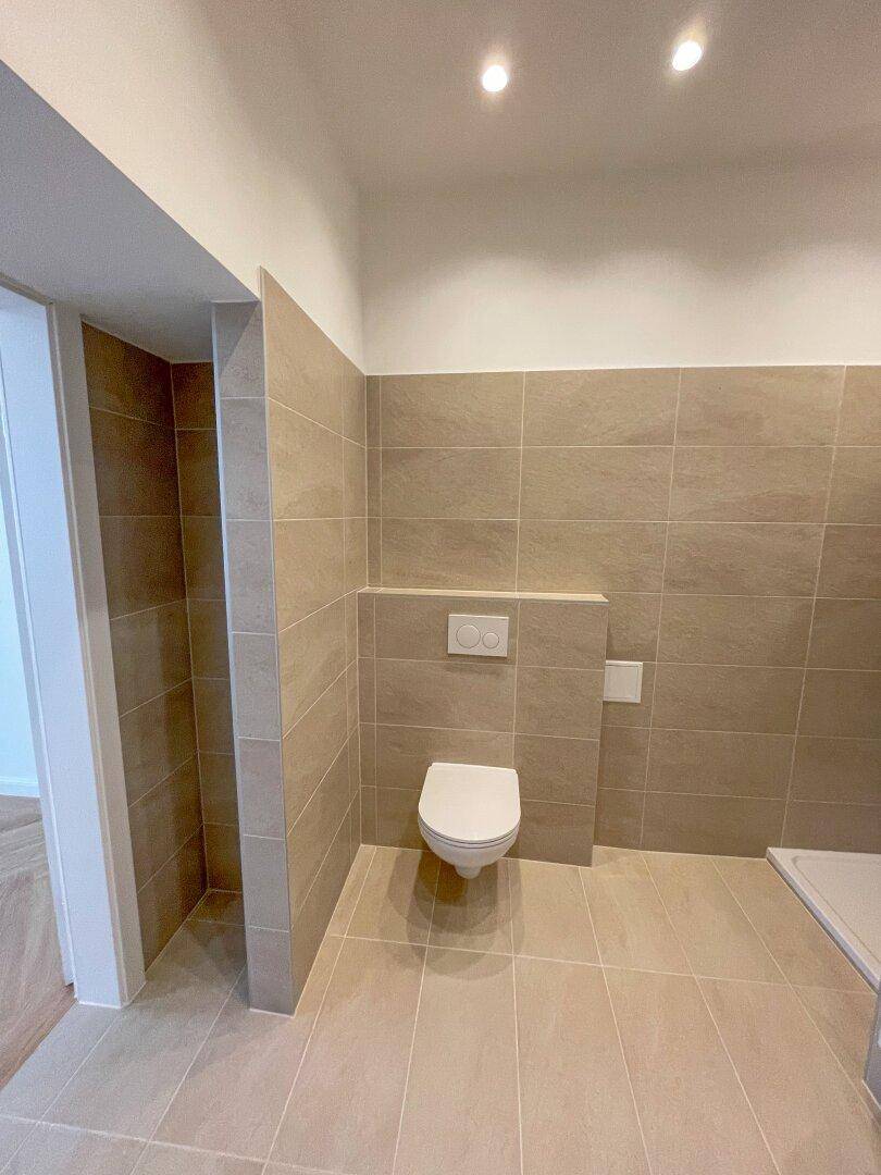 Zeitgemäß gefliestes Badezimmer mit wandhängendem WC und einem Duschbereich, der auf weitere Ausstattung wartet.