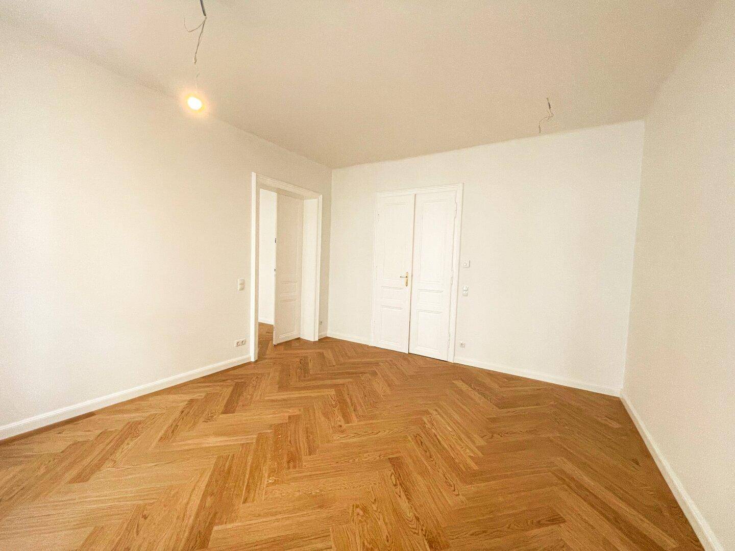 Helles Zimmer mit Fischgrätparkett, zwei Türen und einem offenen Durchgang zu einem angrenzenden Bereich.
