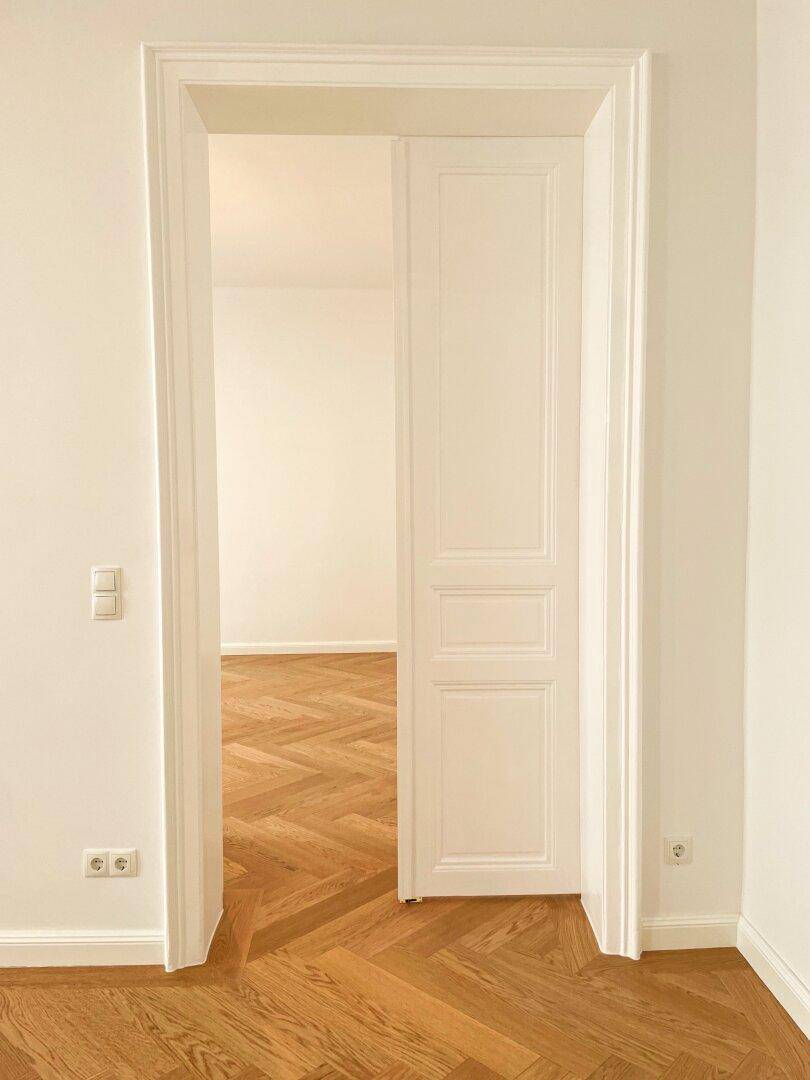 Offene Tür mit elegantem Rahmen, die den Blick auf ein Zimmer mit klassischem Fischgrätparkett freigibt.
