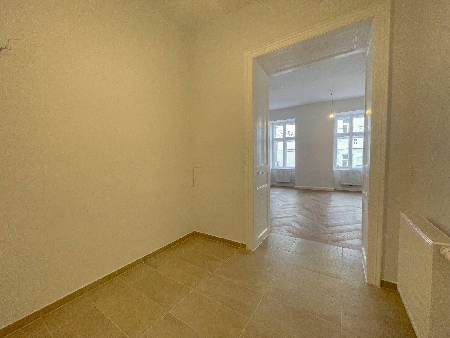 Flur mit Fliesenboden und Heizkörper, der zu einem Zimmer mit elegantem Fischgrätparkett führt.