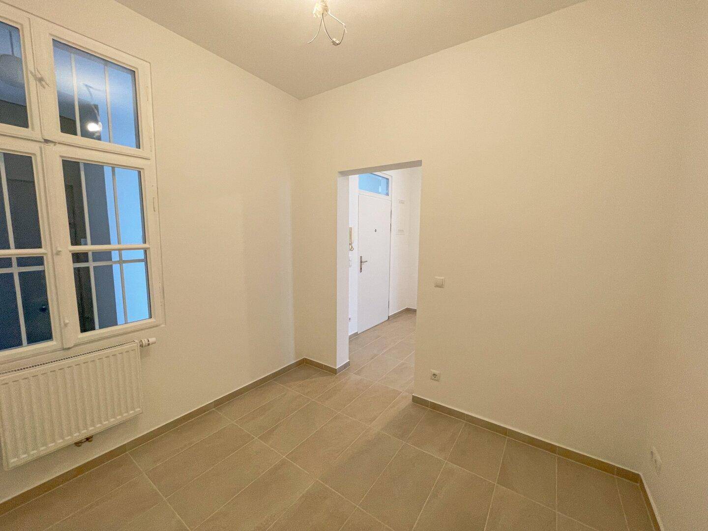 Heller Küchenraum mit Fliesenboden, Fenster und vorbereiteten Anschlüssen für die Kücheninstallation.