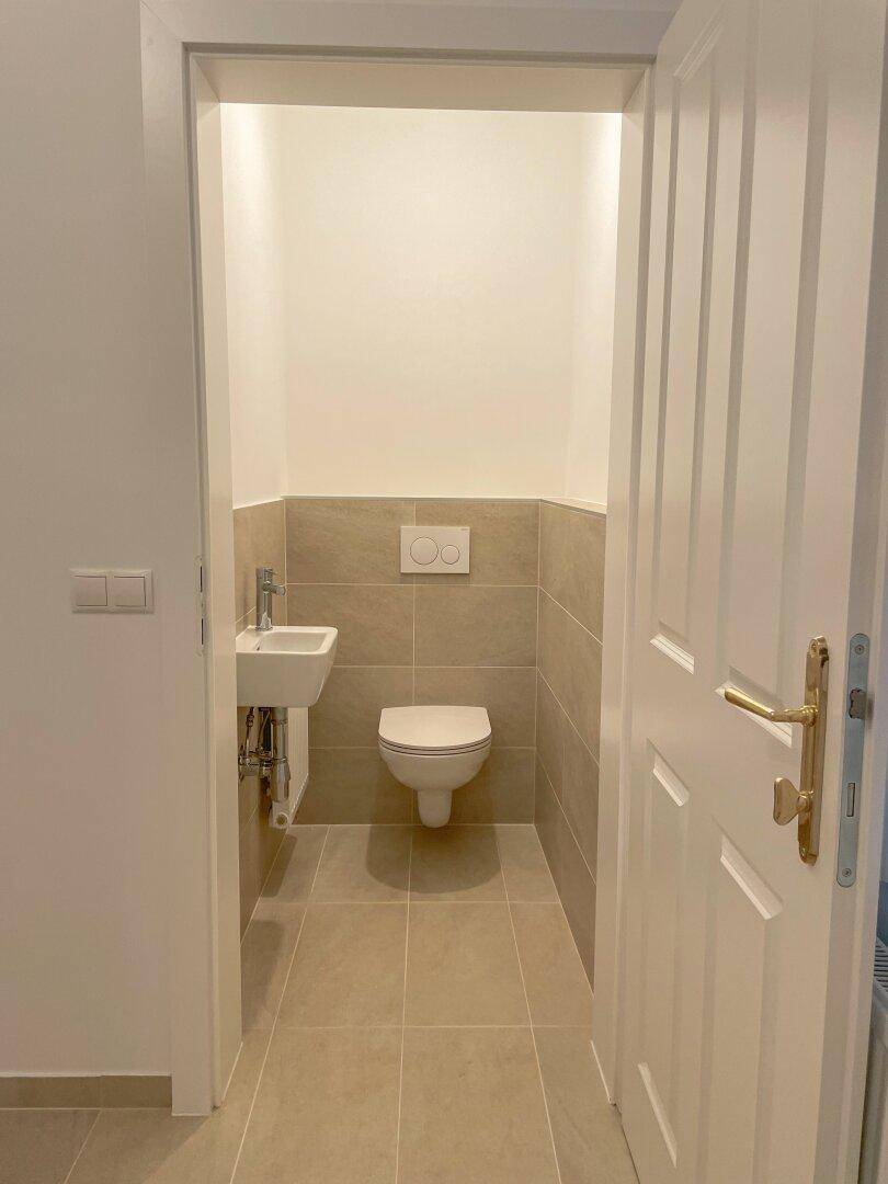 Modernes Gäste-WC mit Fliesenboden, wandhängendem WC und kleinem Waschbecken, funktional und hell.
