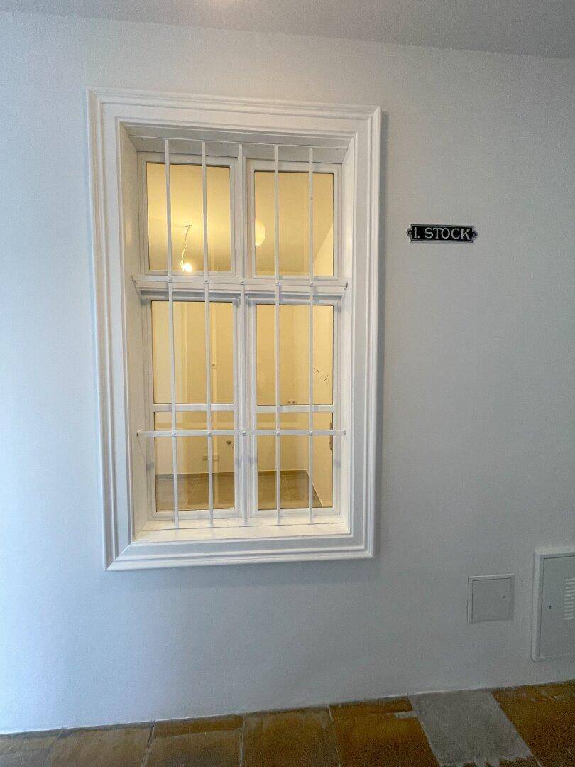Fenster im Treppenhaus mit Gitterstäben und Stockwerksanzeige, das Licht in den Flur lässt.