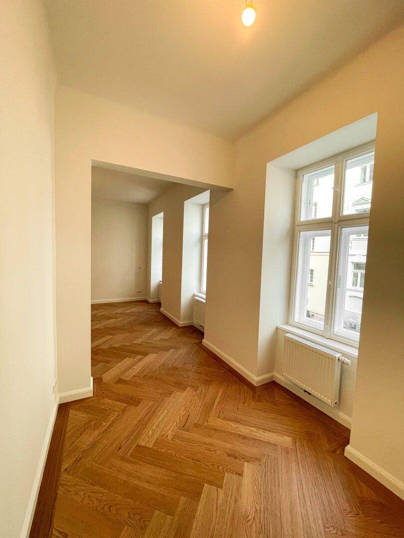 Lichtdurchflutetes Zimmer mit elegantem Fischgrätparkett und mehreren Fenstern, die viel Tageslicht hereinlassen.