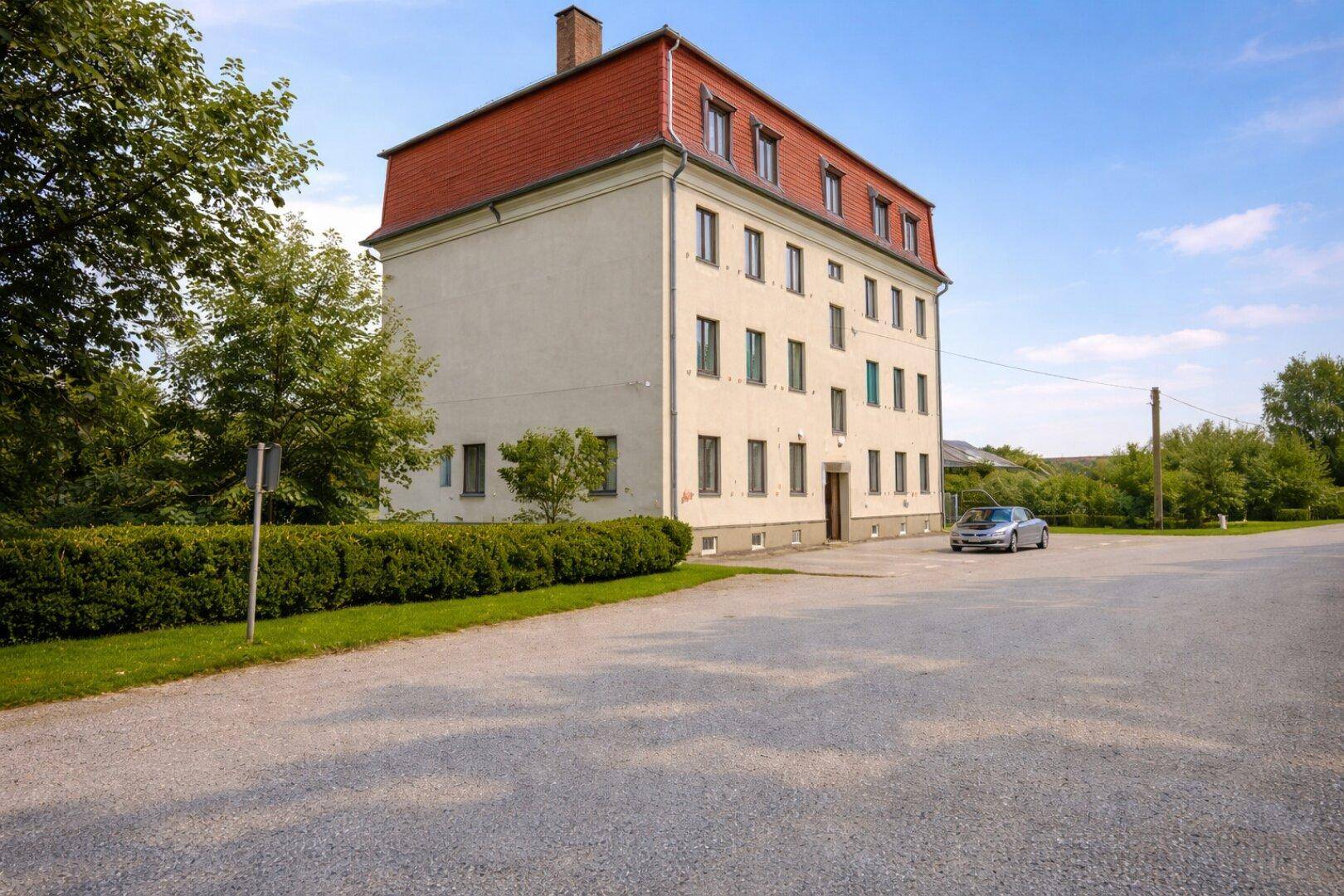 Seitenansicht des Gebäudes mit gepflegter Hecke, Bäumen und einem Parkplatzbereich im Freien.