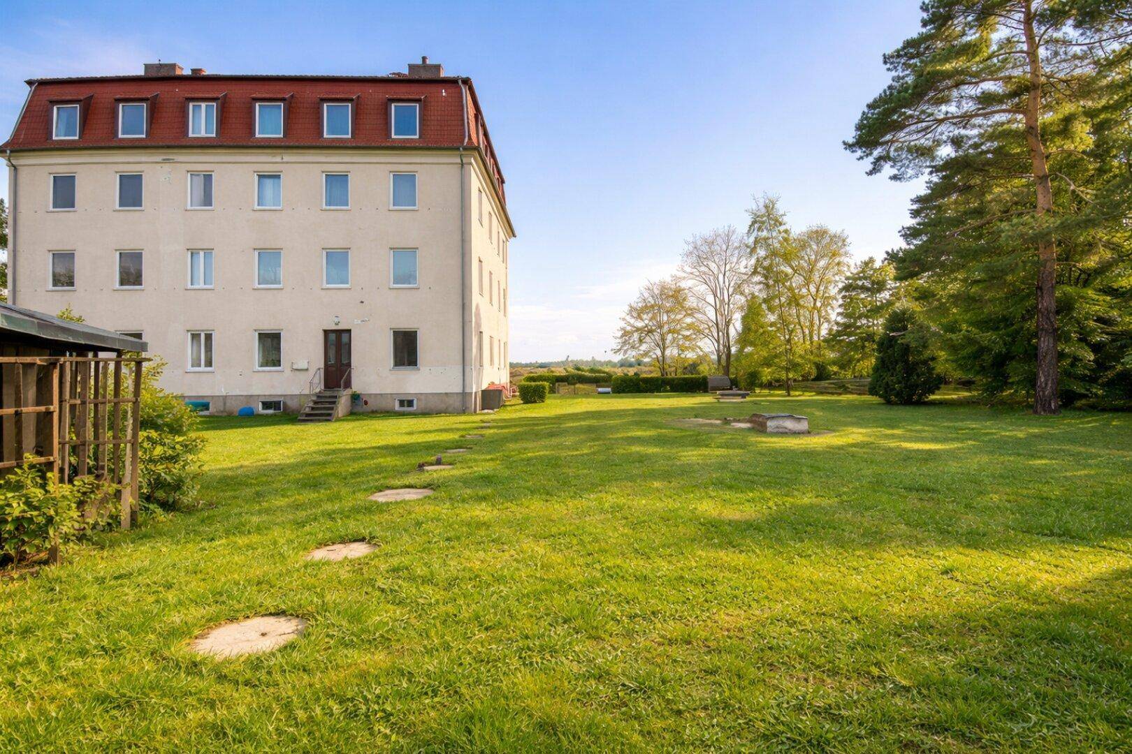 Die Rückseite des Gebäudes mit einem weitläufigen, grünen Gartenbereich und hohen Bäumen.