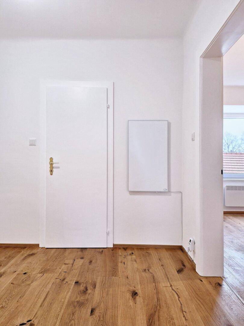 Weiße Wand mit Tür und einem an der Wand montierten Infrarotheizkörper über Holzboden.