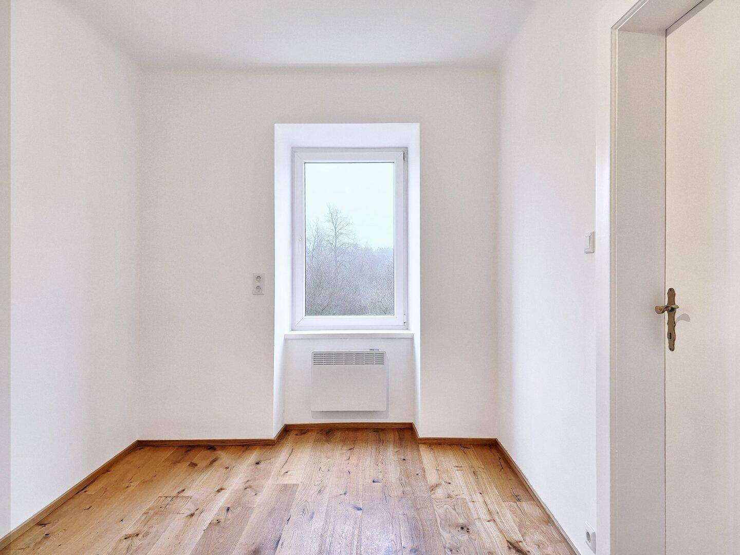 Heller Raum mit Holzboden und einem Fenster, unter dem sich ein Heizkörper befindet.