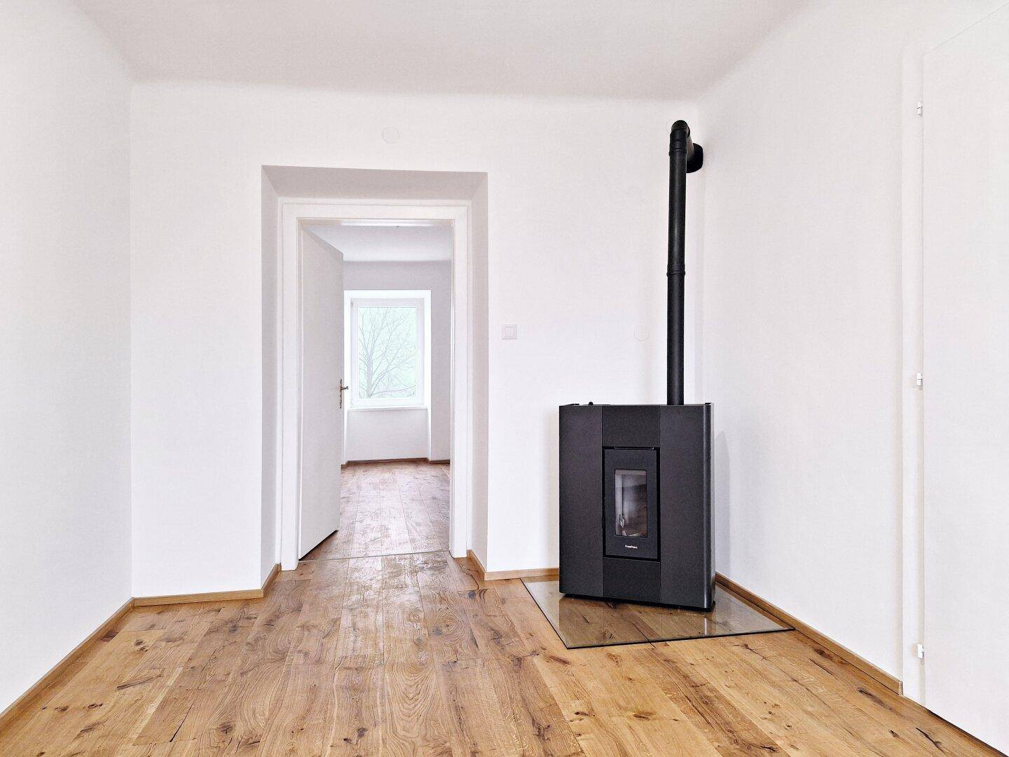 Heller Raum mit Holzboden und modernem Holzofen, der zu einem weiteren Zimmer führt.