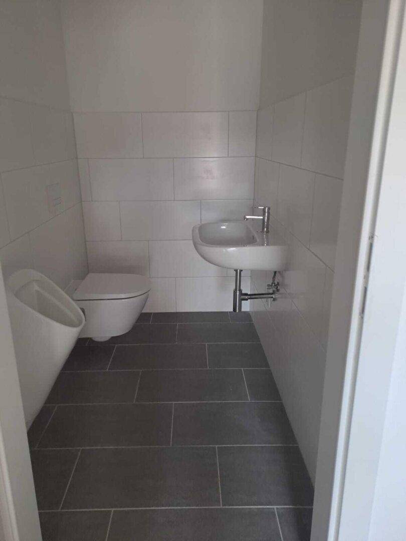 Modernes Gäste-WC mit Urinal, wandhängender Toilette und dunklen Bodenfliesen.