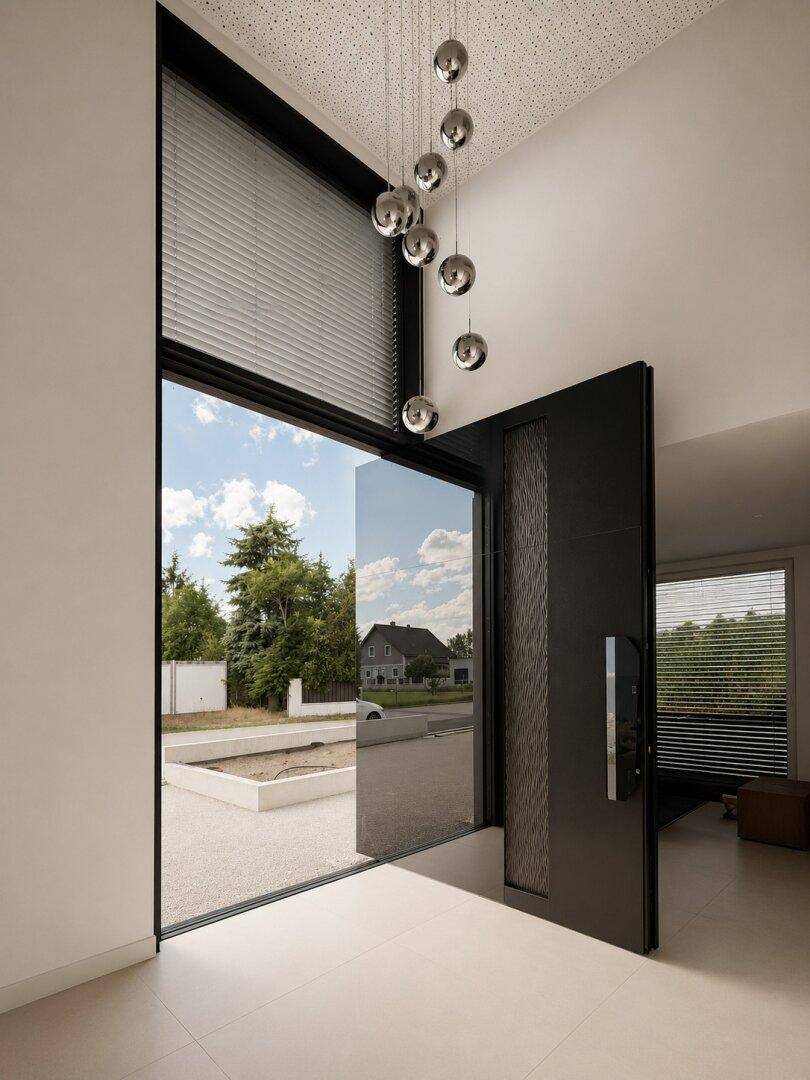 Heller Eingangsbereich mit hoher Decke, moderner Pendelleuchte und großem Glaselement mit Außenblick.