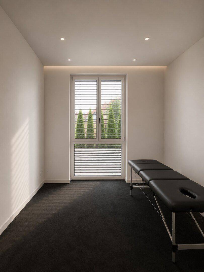 Heller Raum mit dunklem Teppichboden, Fenster und einer Behandlungsliege, ideal für Wellnessanwendungen.