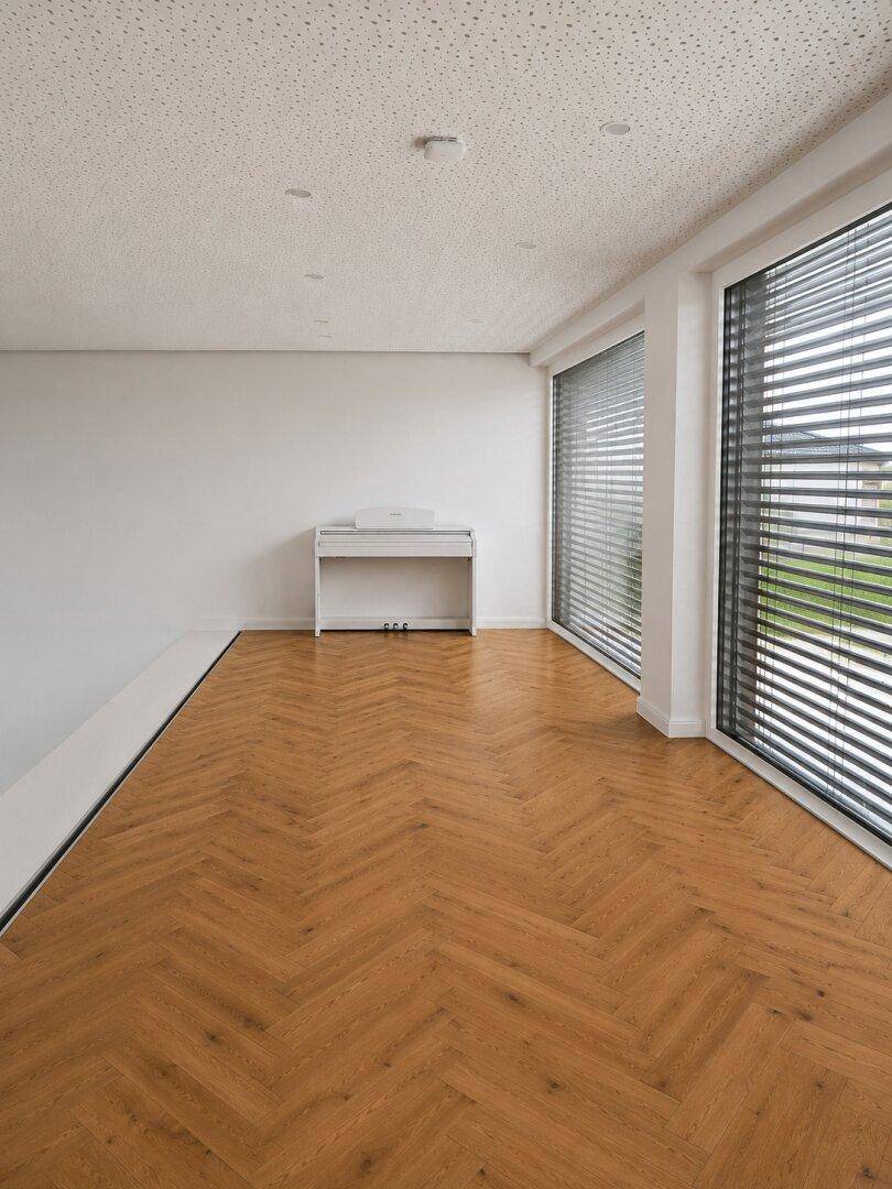 Heller offener Bereich mit Fischgrätparkett, großen Fenstern und einem weißen Klavier, ideal für Musikliebhaber.
