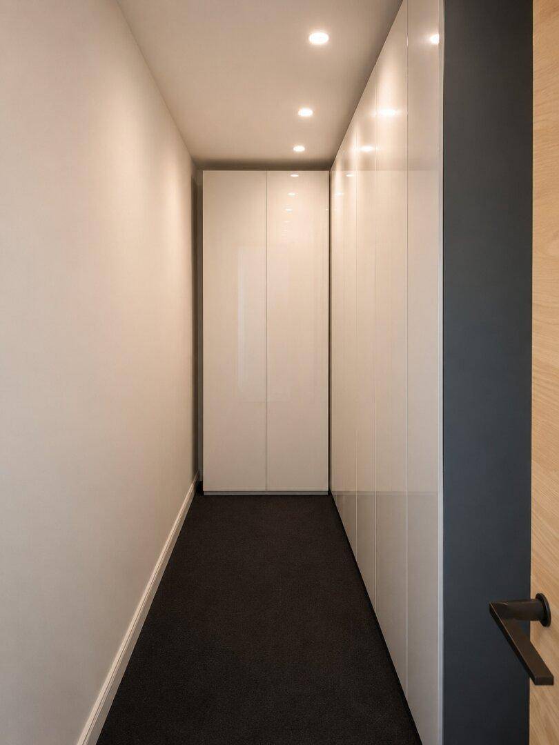 Schmaler Flur mit glänzenden weißen Einbauschränken und dunklem Teppichboden, praktisch und elegant.