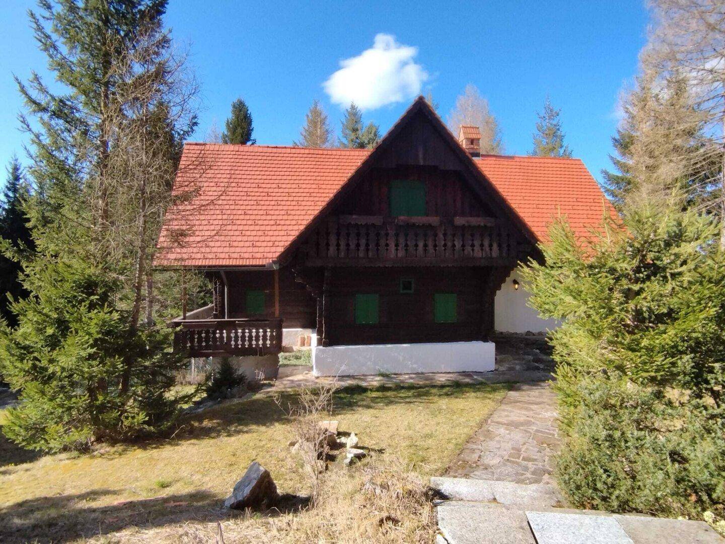 Rustikales Holzhaus mit rotem Ziegeldach, umgeben von Bäumen unter blauem Himmel.