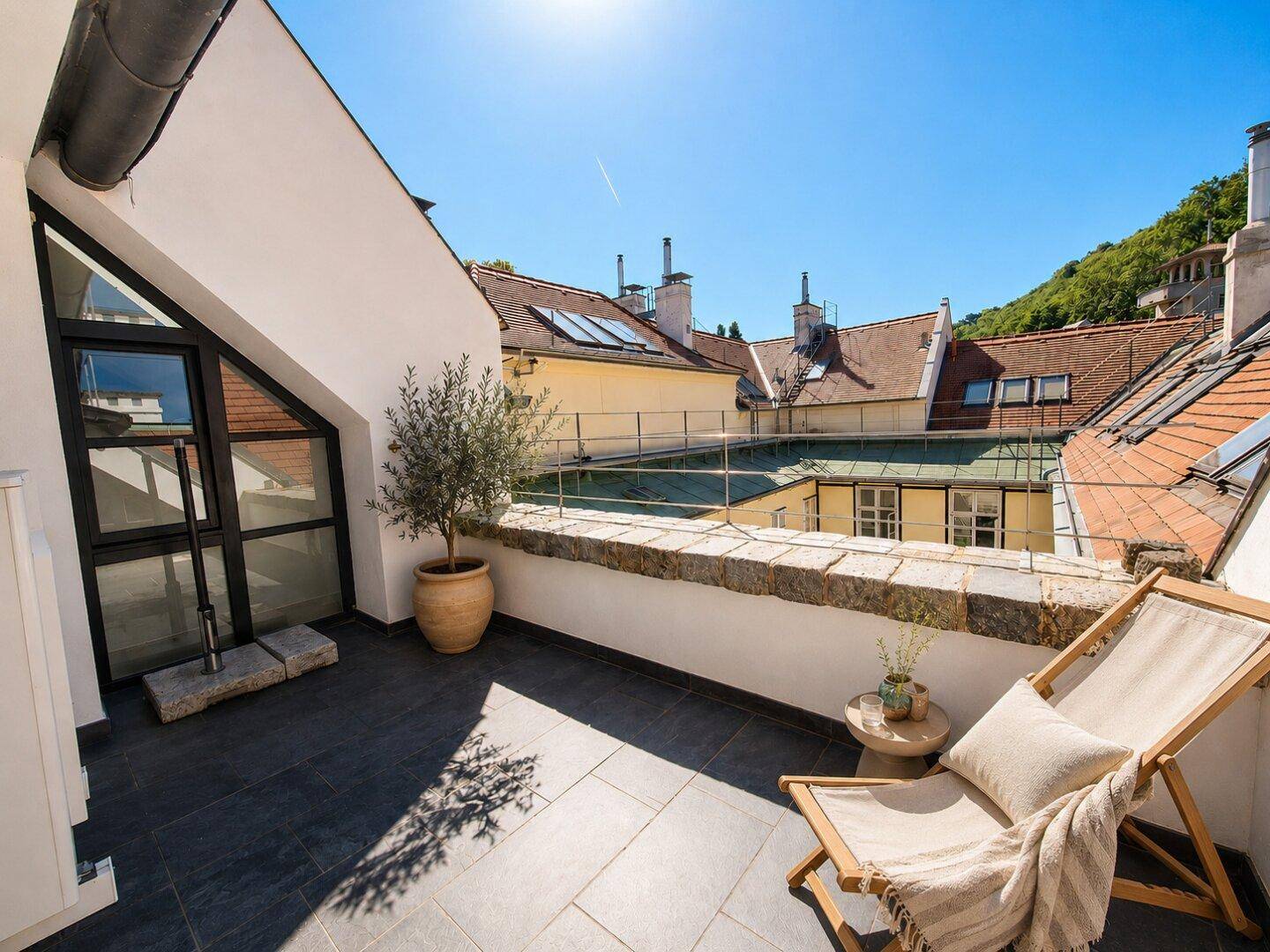 Sonnenterrasse mit Liegestuhl und Blick über die Dächer der Stadt und grüne Hügel.