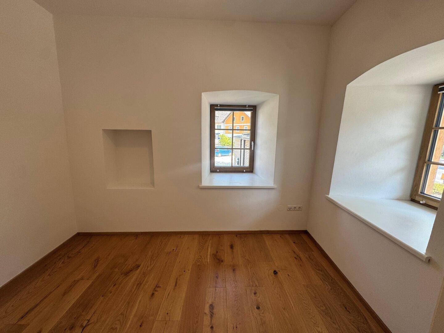 Kleiner, heller Raum mit Holzboden, einem Fenster und einer Wandnische.