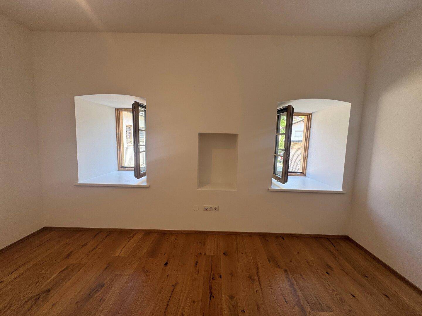 Heller Raum mit zwei kleinen Fenstern und einer Nische in der Wand, ausgestattet mit Holzboden.