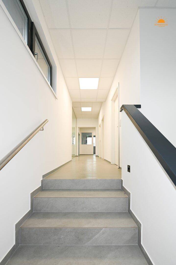 Langer, heller Flur mit grauen Fliesen und abgehängter Decke, der zu weiteren Räumen führt.