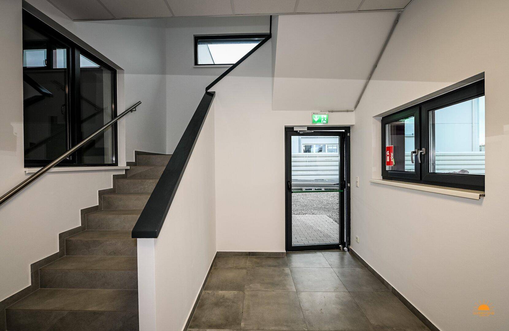 Heller Eingangsbereich mit moderner Treppe, grauen Fliesen und großen Fenstern.