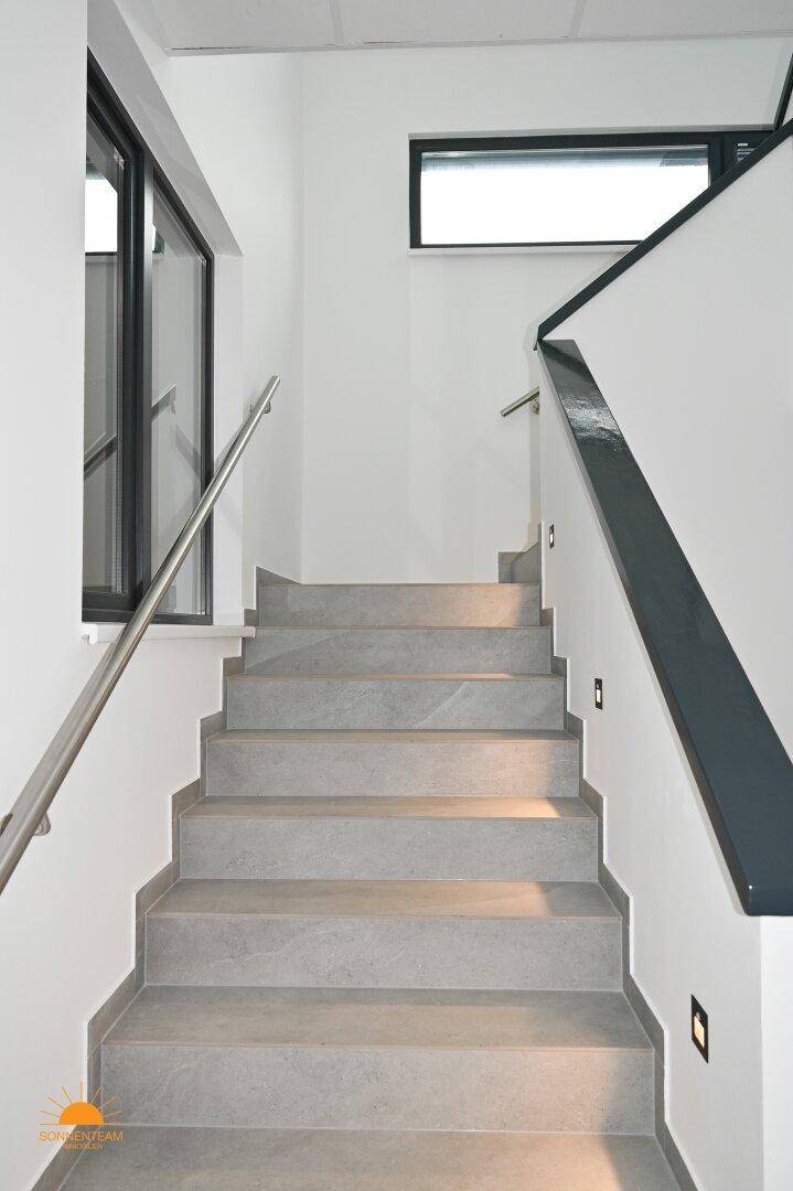 Moderne Treppe mit grauen Stufen und Edelstahlgeländer, beleuchtet durch integrierte Wandleuchten.