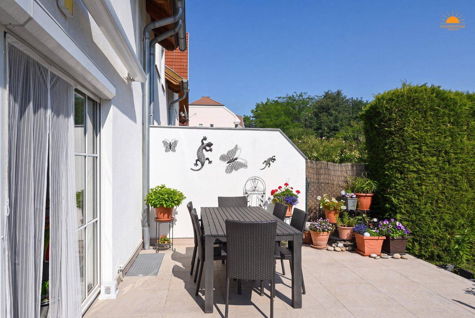 Sonniger Terrassenbereich mit Gartenmöbeln, Pflanzen und hoher Hecke für Privatsphäre.