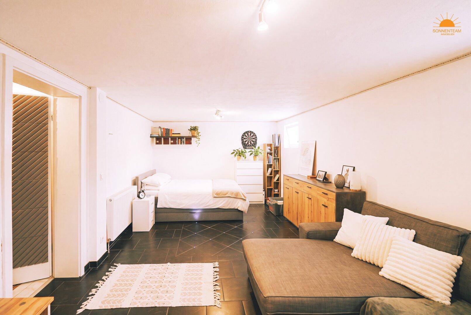 Ausgebauter Kellerraum mit Schlafbereich, Sofa und dunklem Fliesenboden.