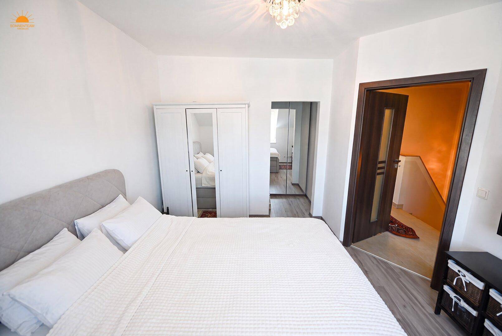 Schlafzimmer mit Doppelbett, weißem Kleiderschrank und Zugang zu einem weiteren Raum.