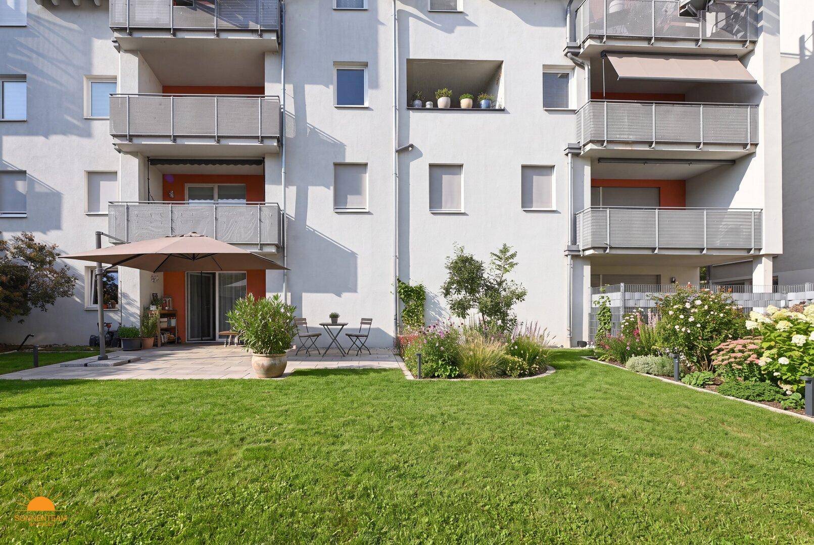Sonniger Gartenbereich mit kleiner Terrasse, Sonnenschirm und Sitzgelegenheiten vor der modernen Fassade.