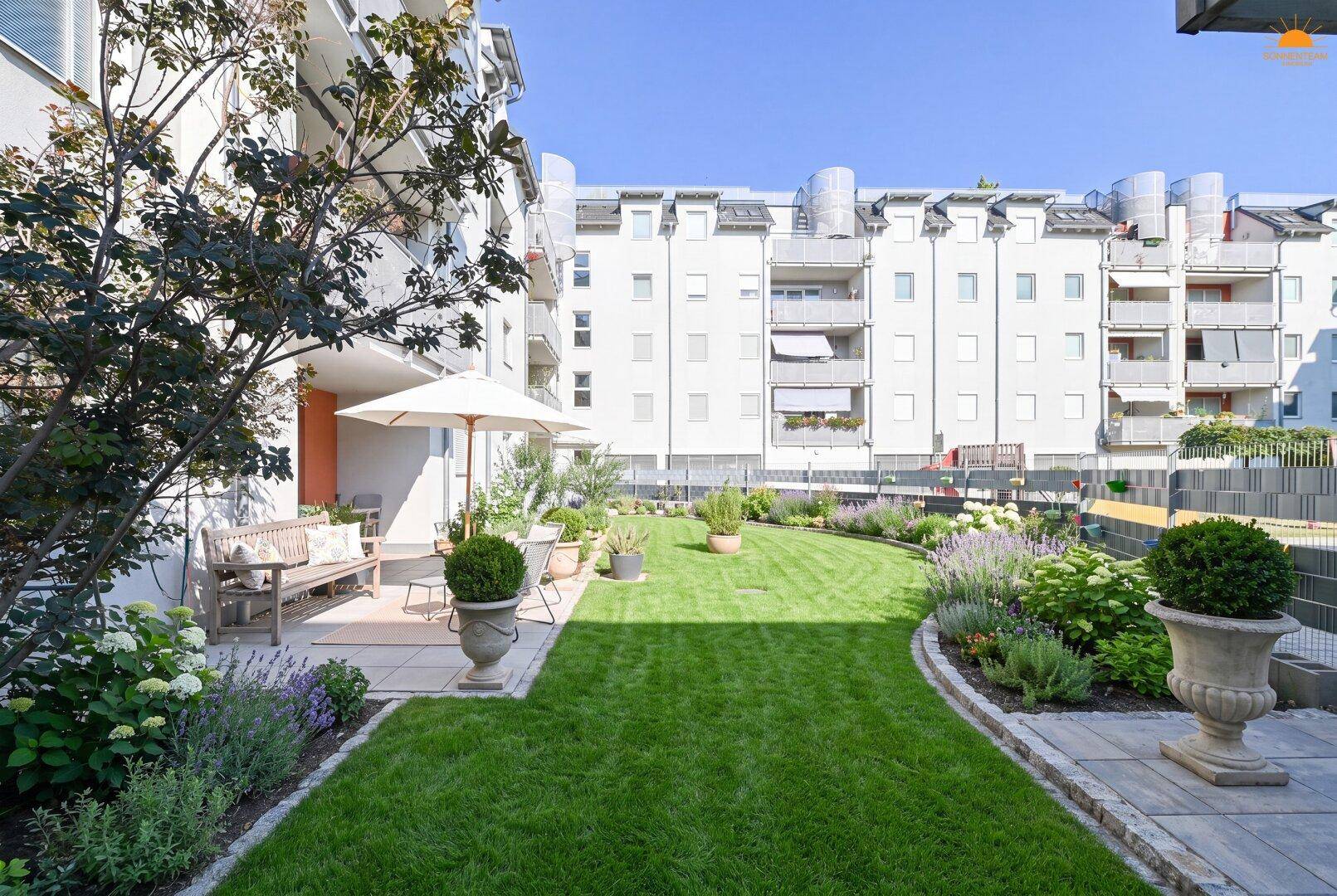 Weitläufiger Gemeinschaftsgarten mit Rasen, Blumen und einer Terrasse im Innenhof der Wohnanlage.