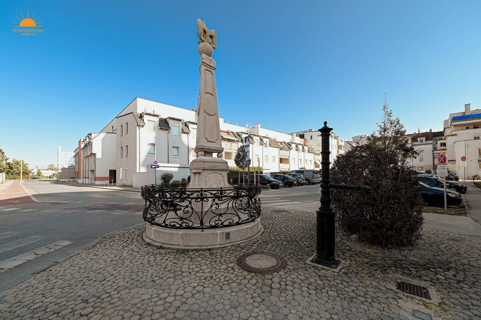 Historischer Brunnen auf einem gepflasterten Platz in der ruhigen Wohngegend.