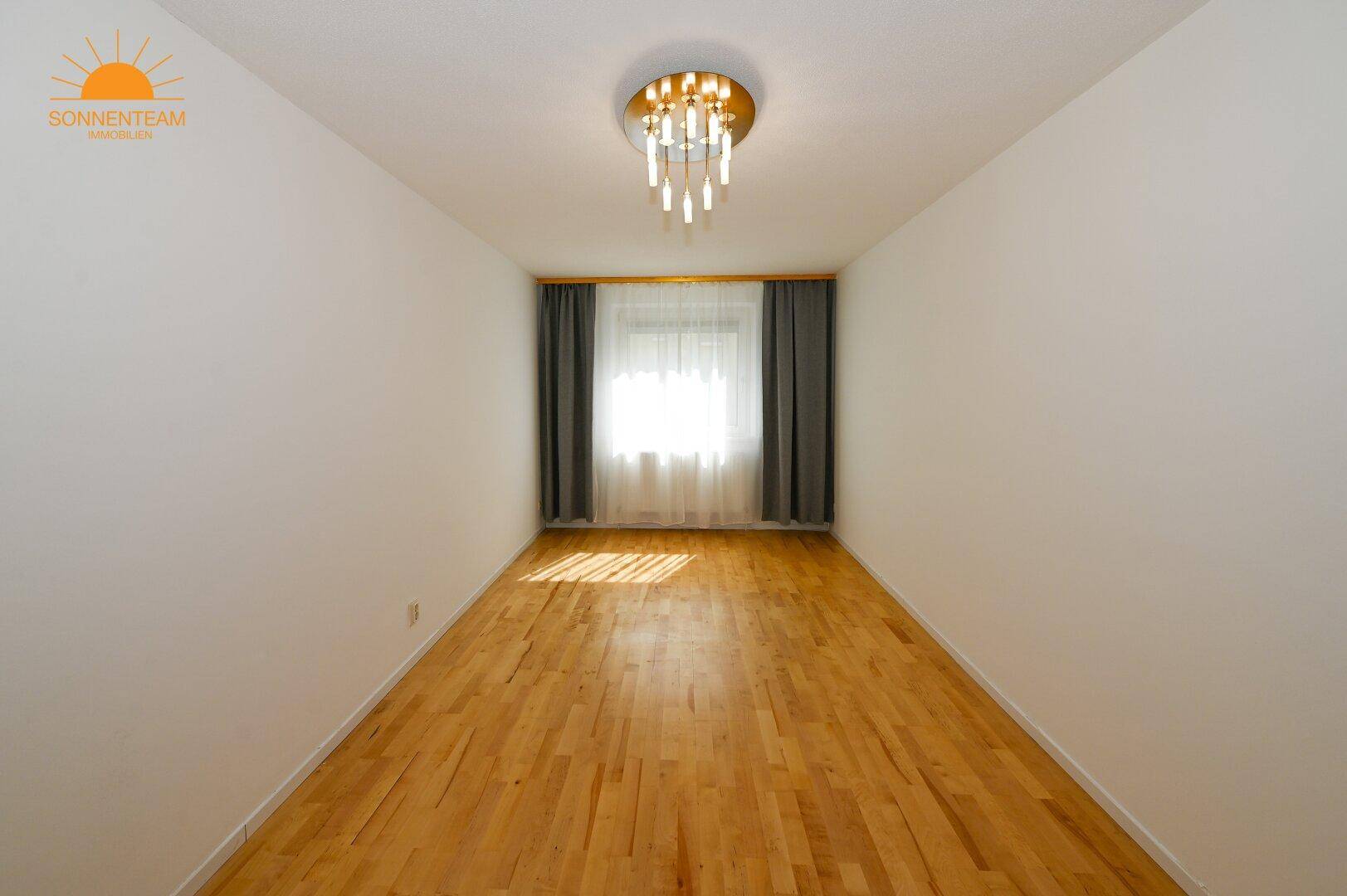 Leerer Raum mit hellem Parkettboden, großem Fenster und moderner Deckenleuchte.