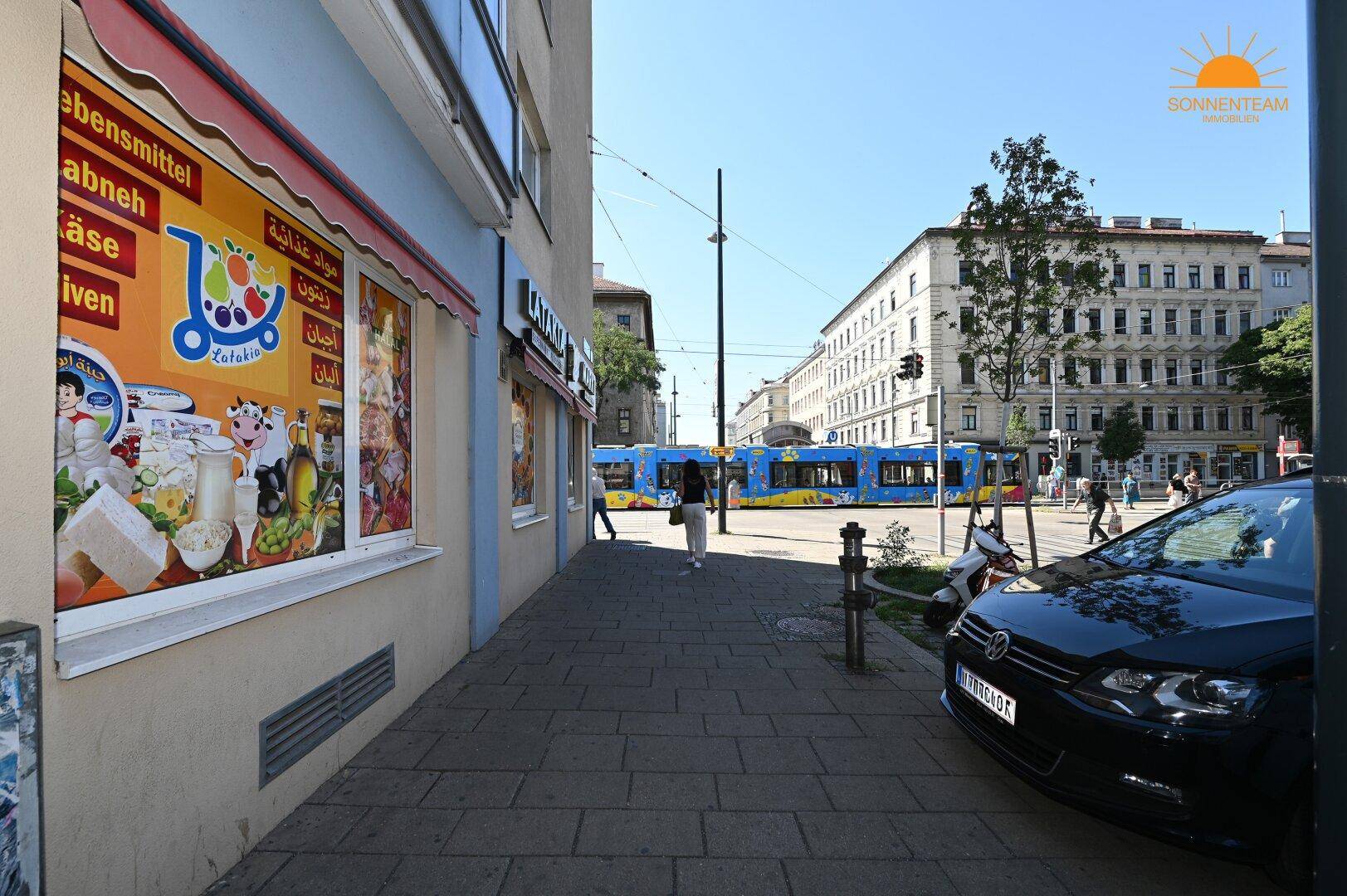 Belebte Straßenszene mit Geschäften, Passanten, einem geparkten Auto und einer Straßenbahn im Hintergrund.