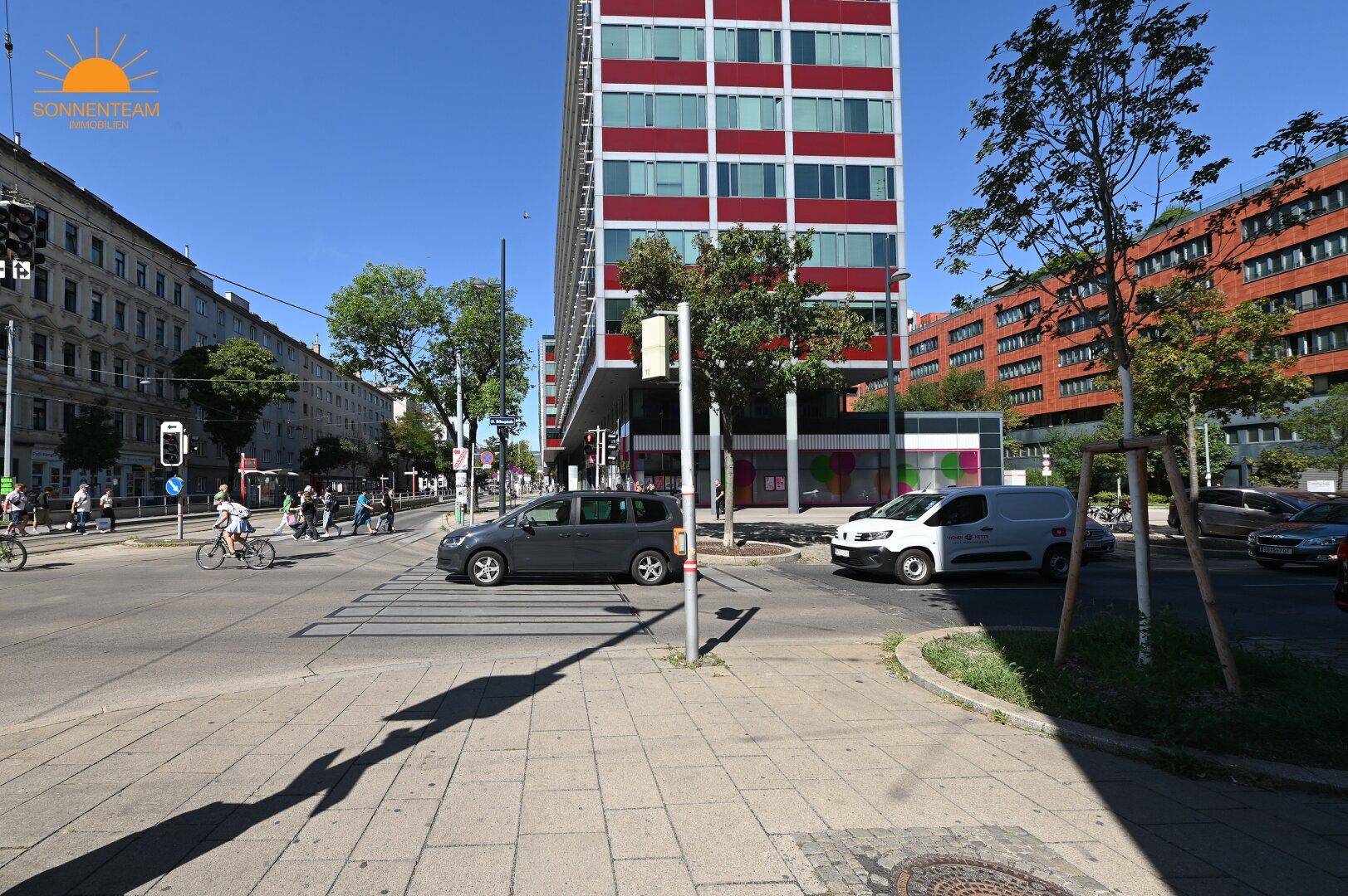 Städtische Kreuzung mit Verkehr, Fußgängern und modernen Gebäuden unter strahlend blauem Himmel.