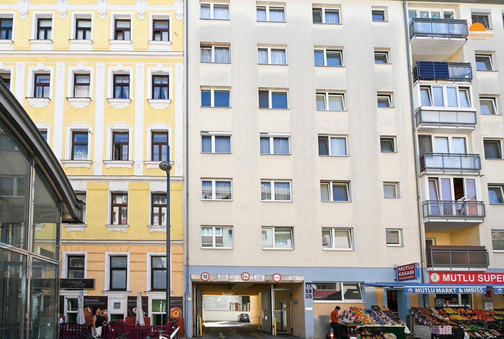 Fassade eines Mehrfamilienhauses mit Balkonen, Geschäften im Erdgeschoss und einer Tiefgarageneinfahrt.