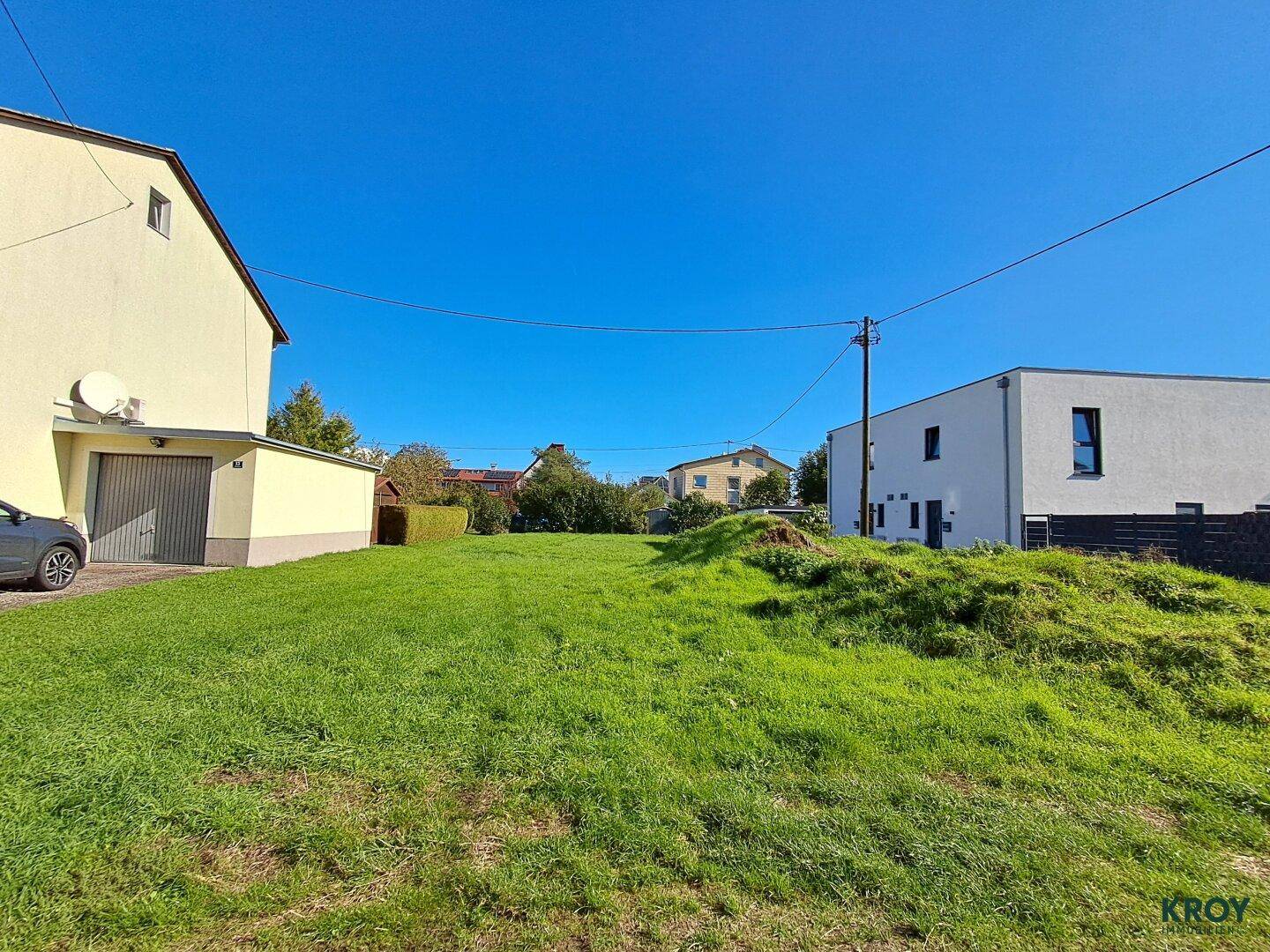 Grüne Wiese eines Baugrundstücks mit angrenzenden Wohnhäusern unter blauem Himmel.