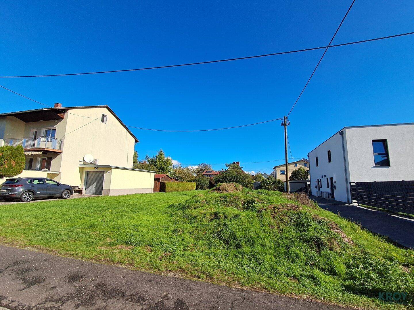Sonniges Baugrundstück mit Grasfläche und angrenzender Straße sowie Wohnhäusern.