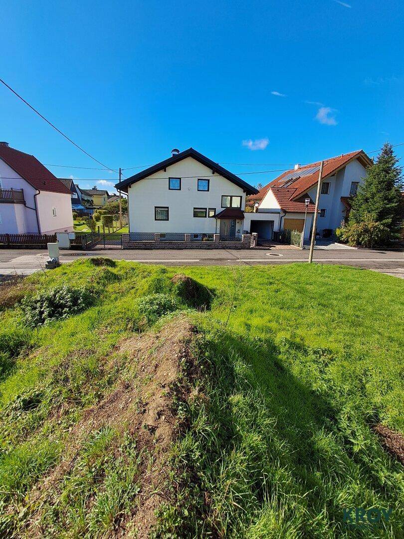 Grünes Baugrundstück mit leichter Erhebung und Wohnhäusern auf der gegenüberliegenden Straßenseite.