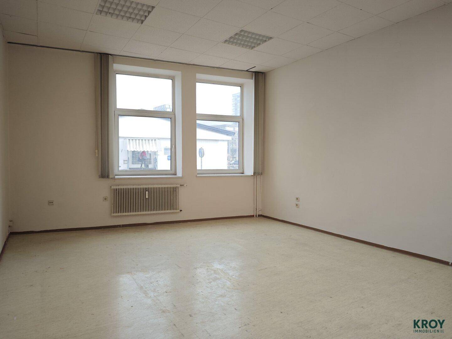 Heller, leerer Raum mit zwei Fenstern und einer abgehängten Decke.