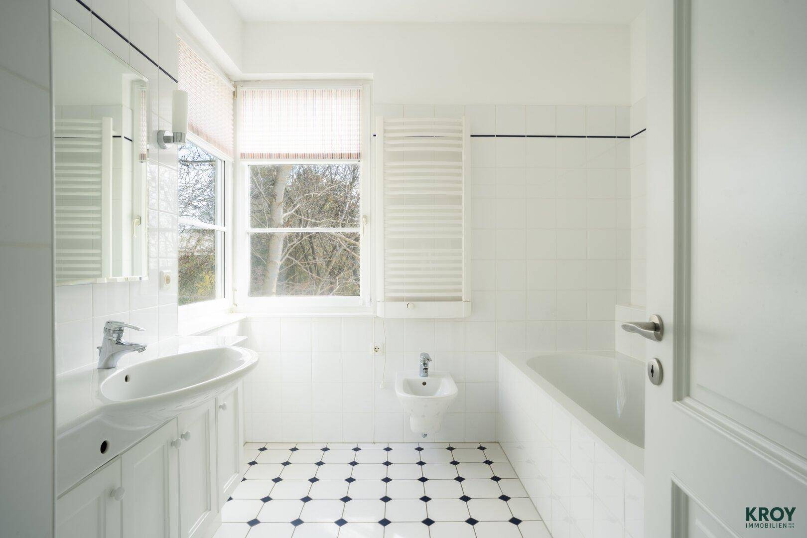 Helles Badezimmer mit Badewanne, Waschbecken, Bidet und Fenster, gefliest in Schwarz-Weiß.