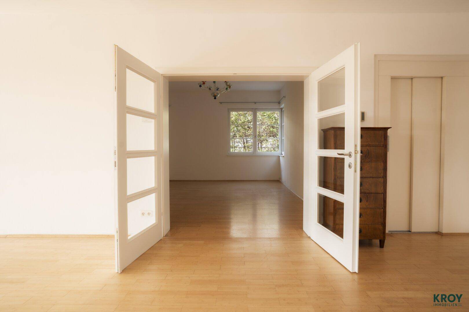 Heller Raum mit Holzboden, Flügeltüren und einem antiken Holzschrank, der vielseitig nutzbar ist.
