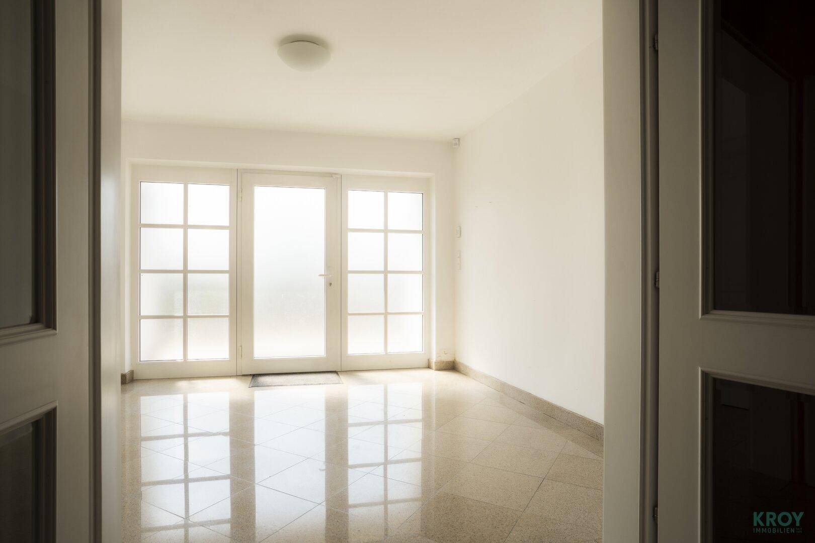 Heller Eingangsbereich mit glänzenden Fliesen und großen Fenstertüren.
