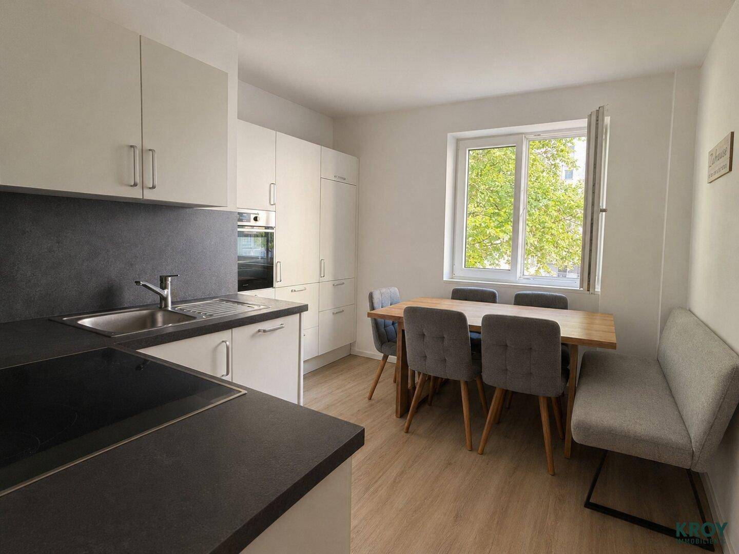 Moderne Küche mit weißen Schränken, dunkler Arbeitsplatte und Essbereich am Fenster.