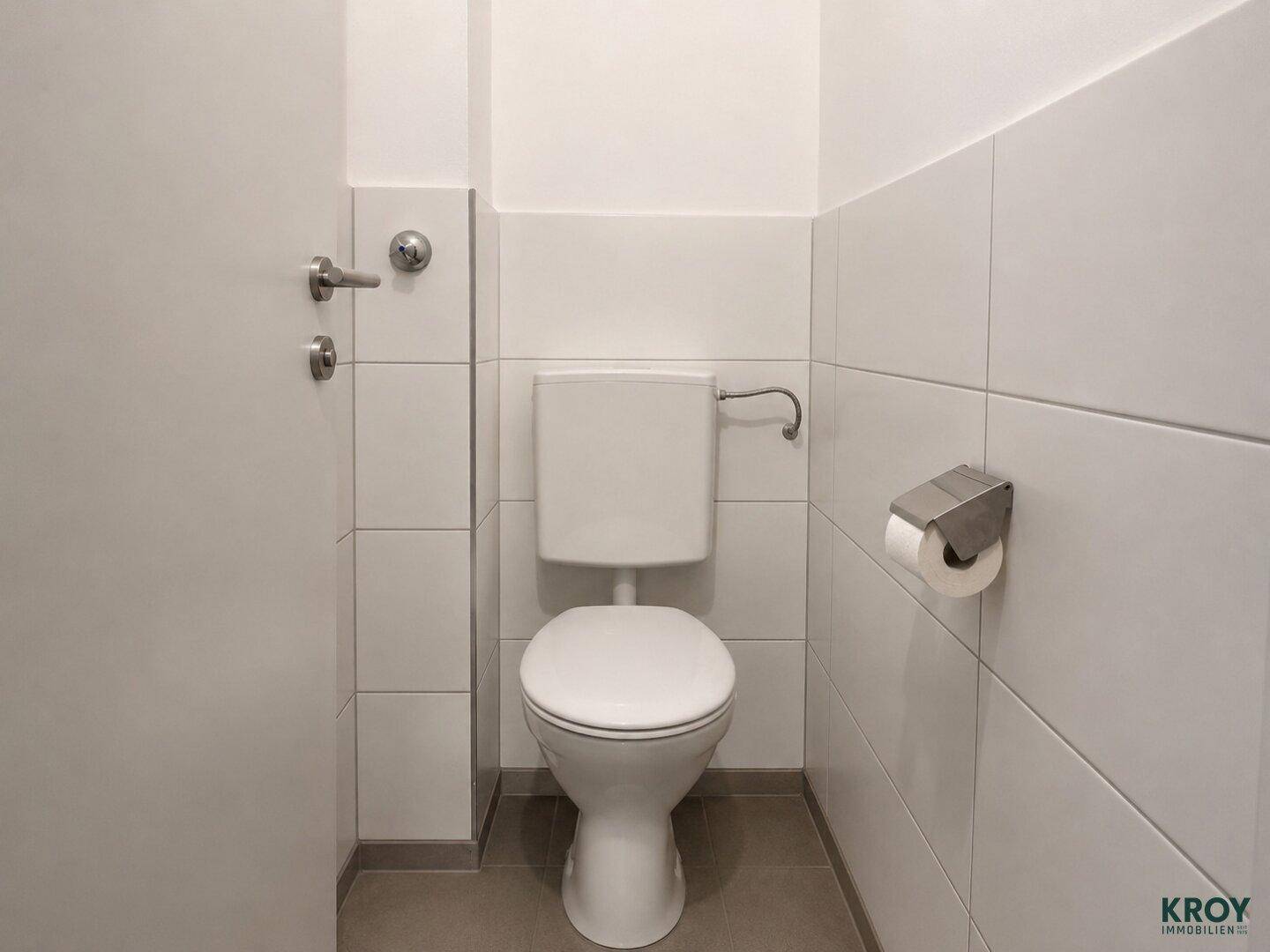 Separates WC mit weißer Toilette und hellen Fliesen an den Wänden.