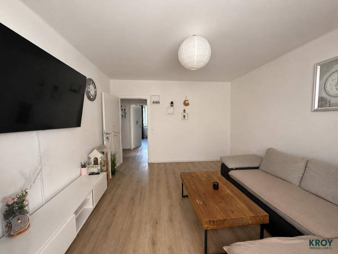 Geräumiges Wohnzimmer mit großem Fernseher an der Wand und hellem Laminatboden.