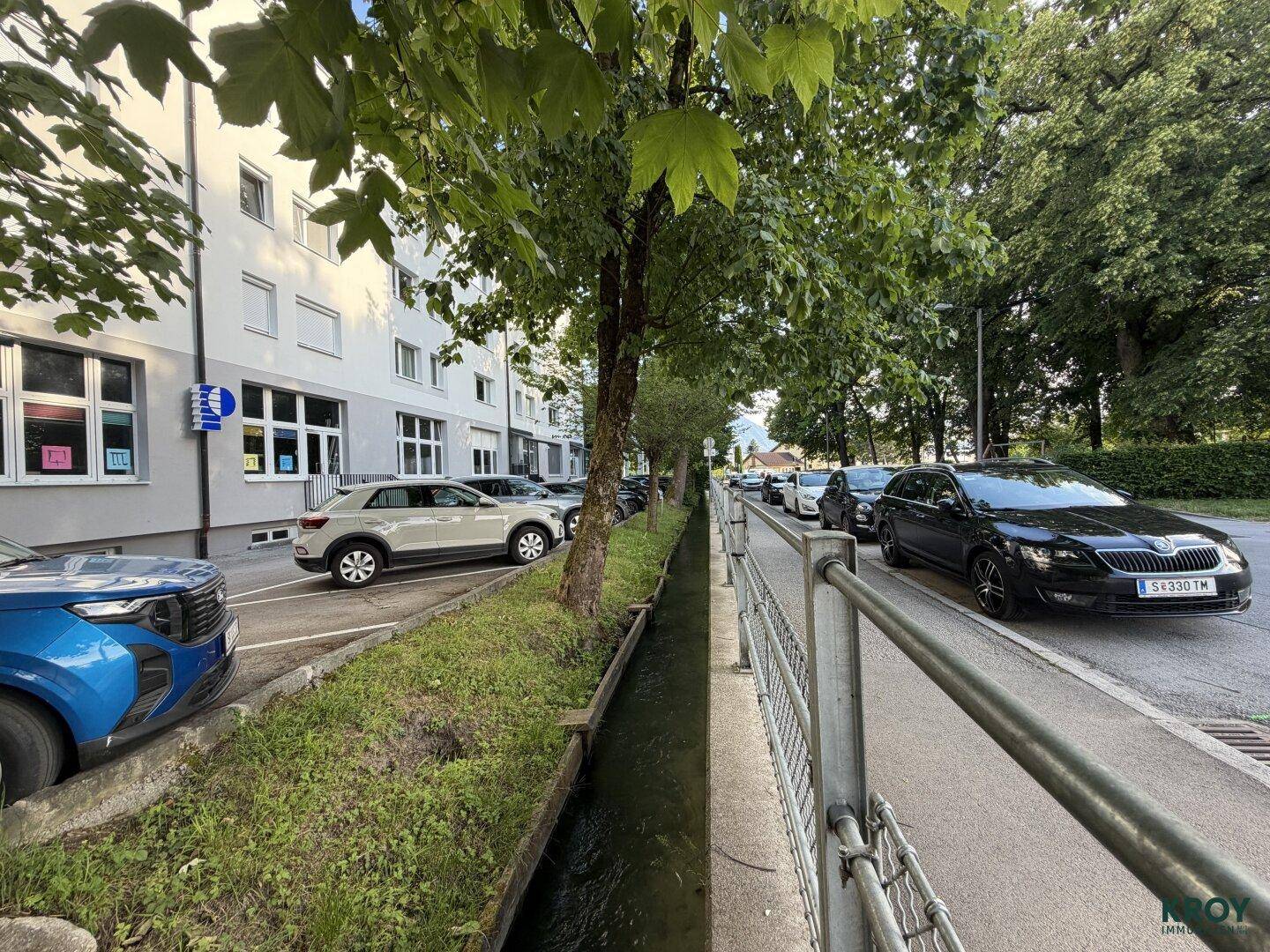 Straße mit parkenden Autos, einem kleinen Kanal und Bäumen vor einem Wohngebäude.