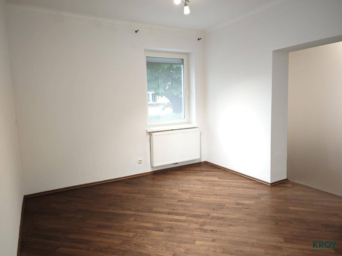 Helles Schlafzimmer mit dunklem Holzboden und einem Fenster mit Blick ins Grüne.